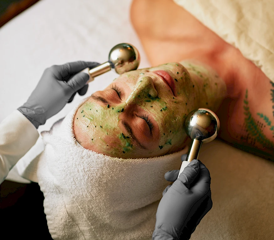 A person receives a green herbal facial treatment with two cupping-like tools; medicated paste on their face while lying with a towel around the head.