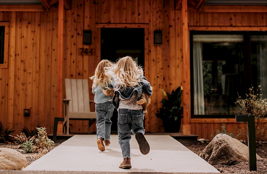 Two children are running toward a wooden house surrounded by trees, under a cloudy sky.