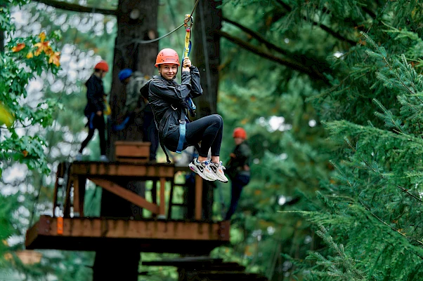 A person wearing a helmet is zip-lining on a platform high among green trees, with others in the background waiting.