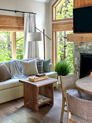 A cozy living room with a sofa, cushions, a fireplace, TV, plants, round wooden table, woven chairs, and large windows.