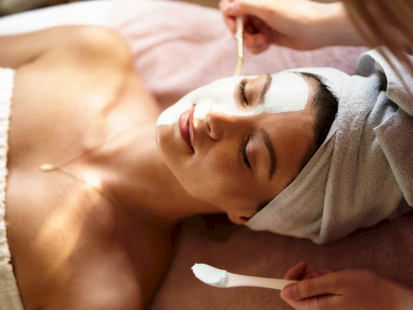 A person is receiving a facial treatment with a mask applied, while lying down with a towel wrapped around their head.