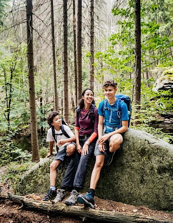 Three people sit on a large rock in a forest, smiling and posing among tall trees and green foliage.